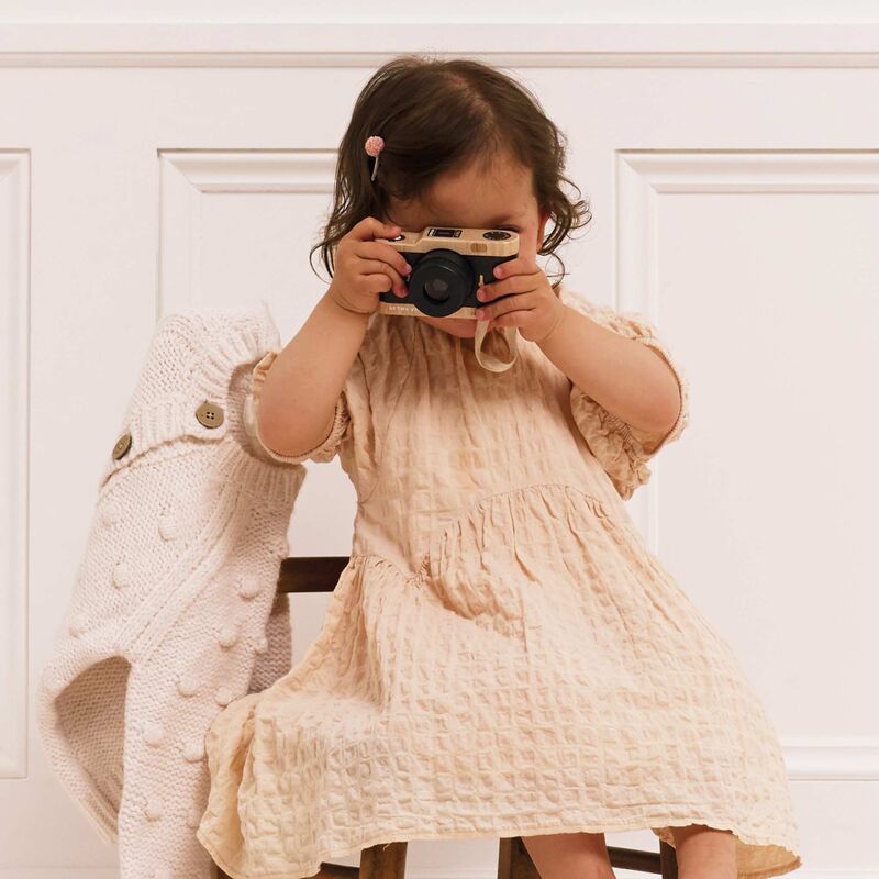 Child holding a camera in front of their face, wearing a beige dress and sweater.